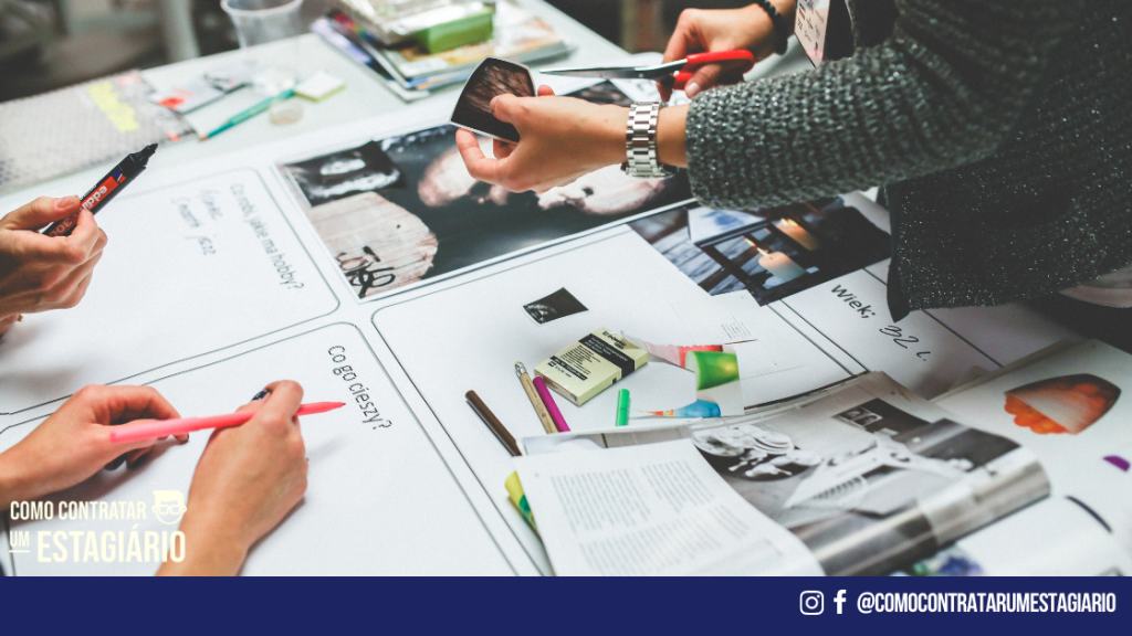 Projetos sendo criados por uma equipe em uma mesa com recortes, cartazes e tesouras. A equipe está construindo qual é o propósito da empresa.