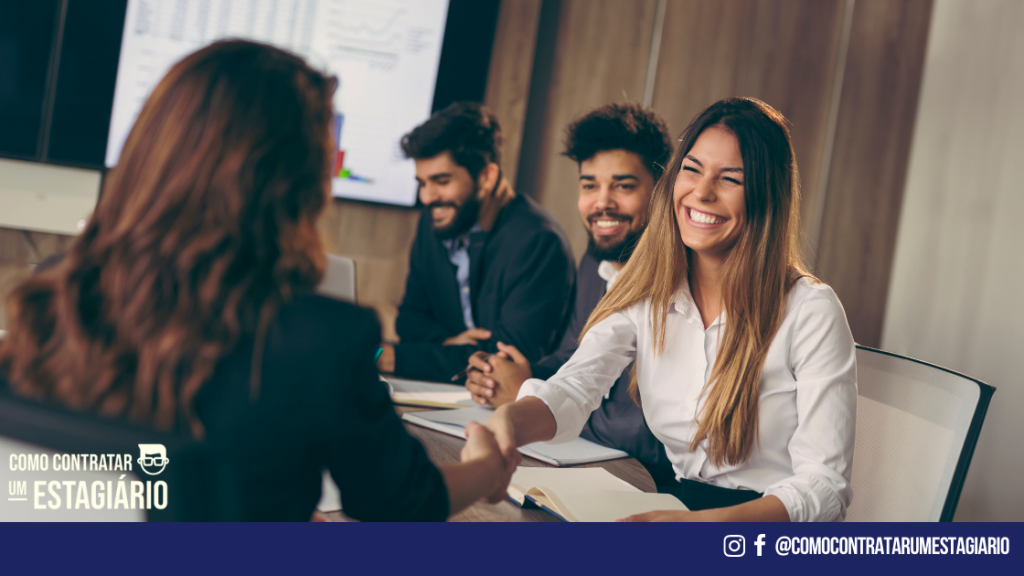 Uma equipe num local de trabalho, sentados em uma mesa. Uma mulher mais jovem está sorrindo e apertando a mão de outra mulher. As duas mulheres estão conversando sobre a possibilidade de fazer dois estágios ao mesmo tempo.