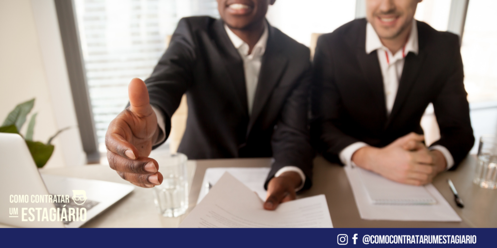 Dois homens sentados vestidos de terno. Um está com papeis na mão, esticando sua mão ao estagiário para fazer um acordo de efetivação do estagiário na empresa.