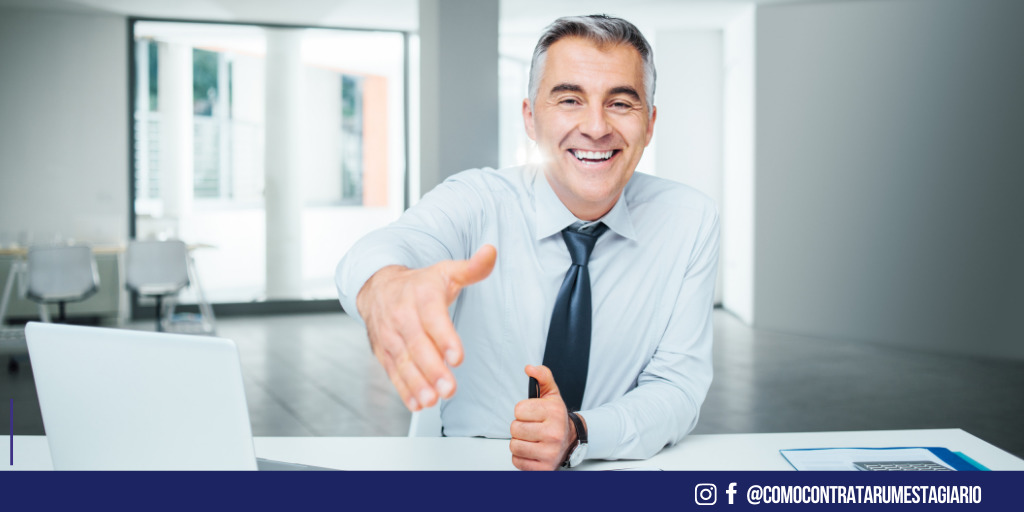 Um homem sorrindo, vestido com roupas sociais. Está sorrindo, estendendo a mão para fechar um contrato de estagiário, selecionado através de uma vaga de estágio.